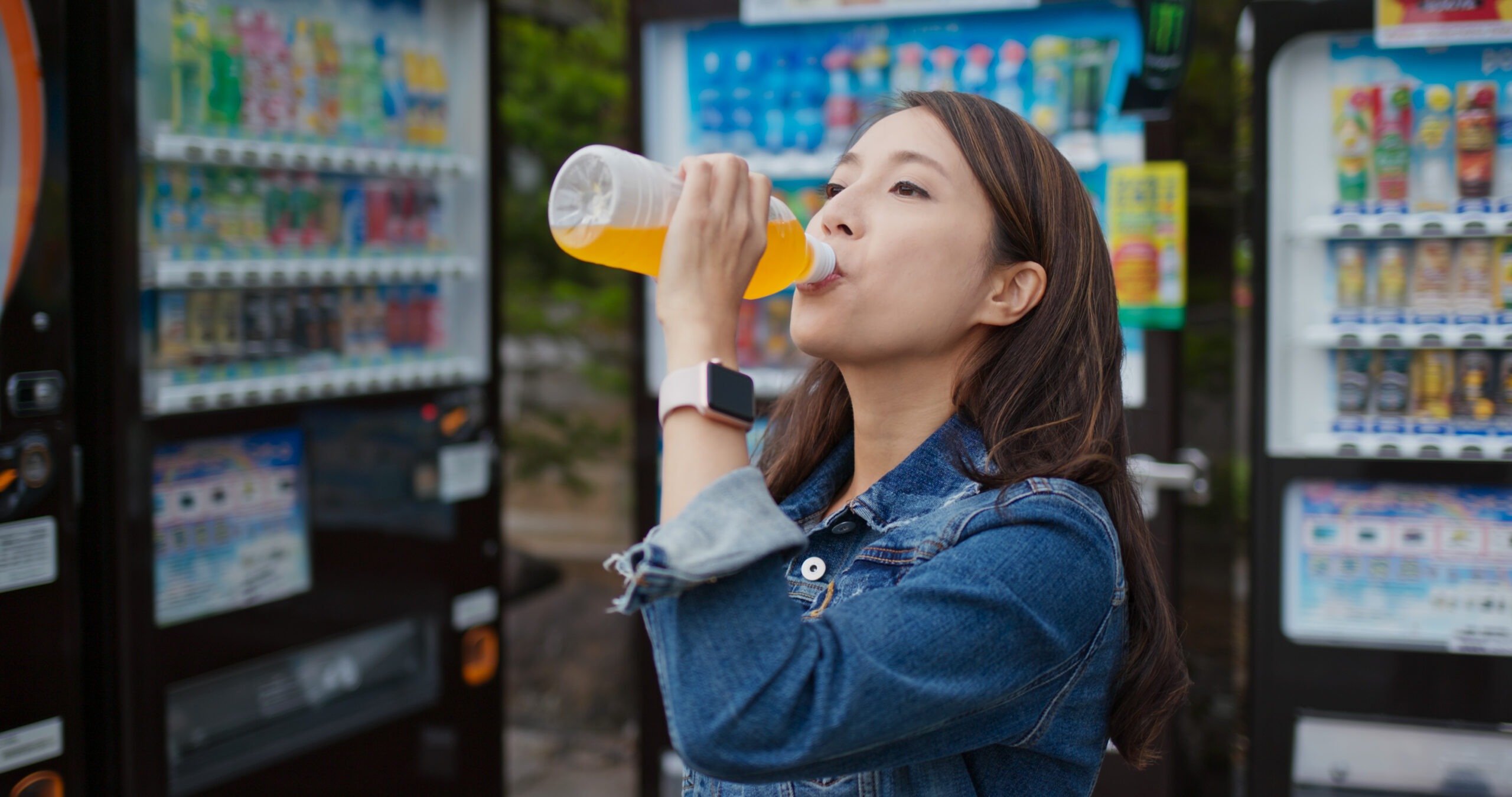 Young woman drinks orange beverage from a bottle in front of vending machines. The scene suggests refreshment and casual outdoor enjoyment.