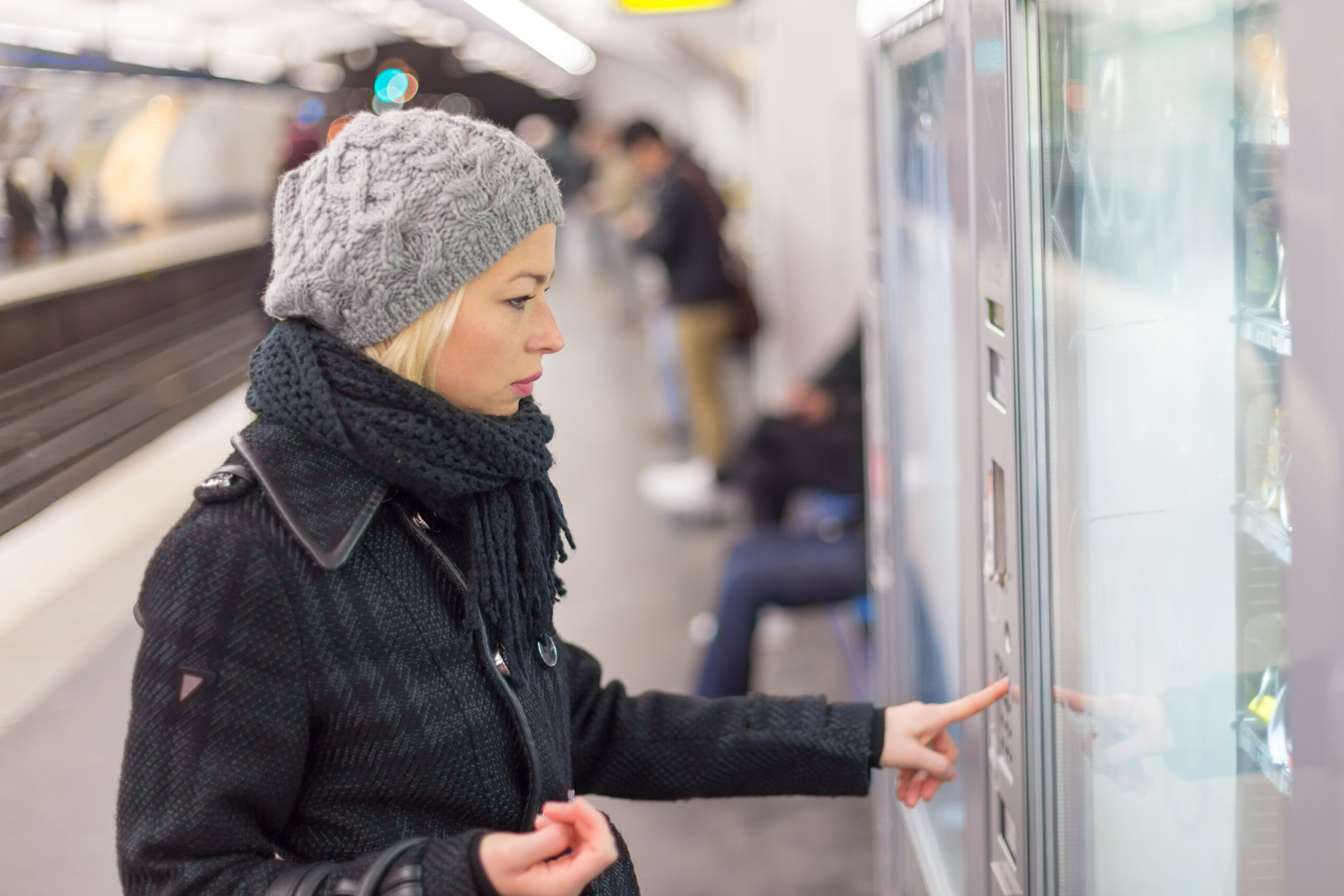 A woman in a black coat and knit hat selects a snack from a vending machine at a subway station, with passengers waiting in the background.