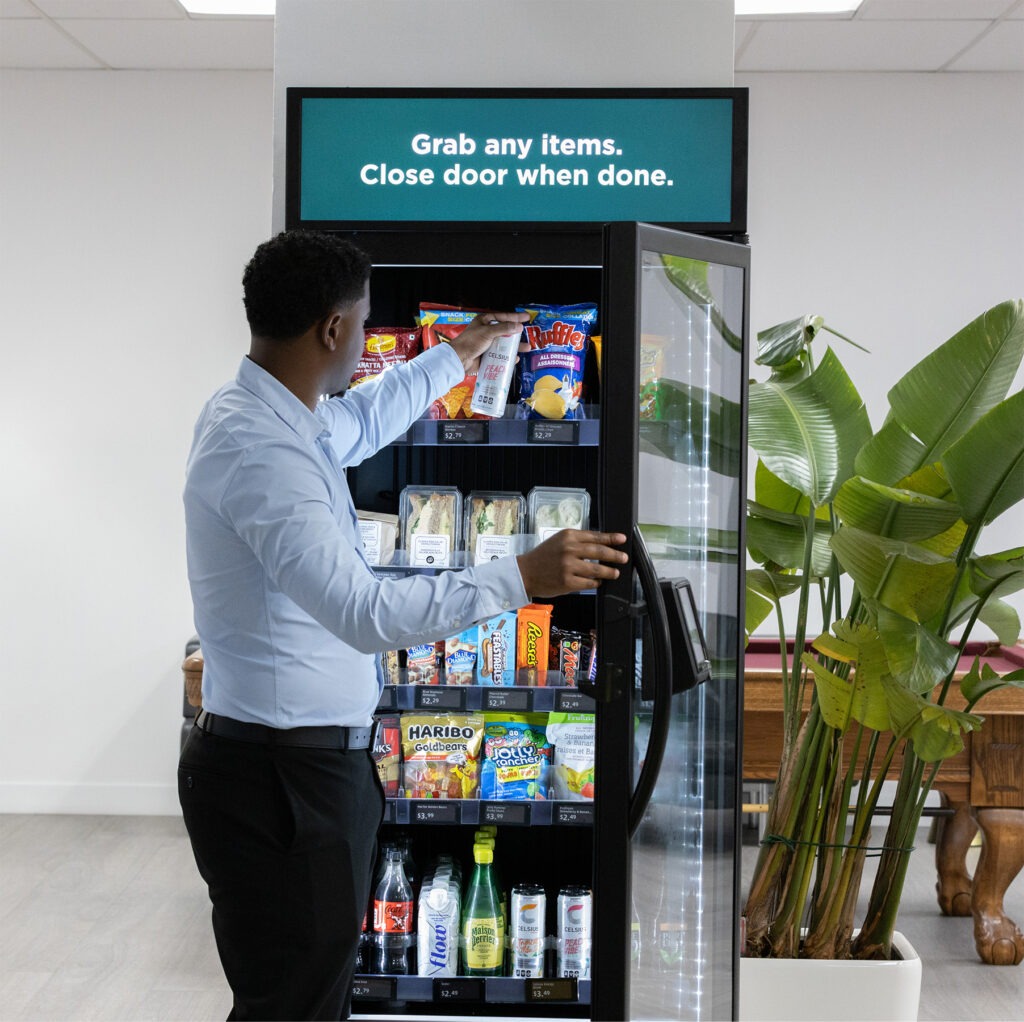 A person in a light blue shirt selects snacks from a vending machine, with a sign above instructing to grab items and close the door when done.