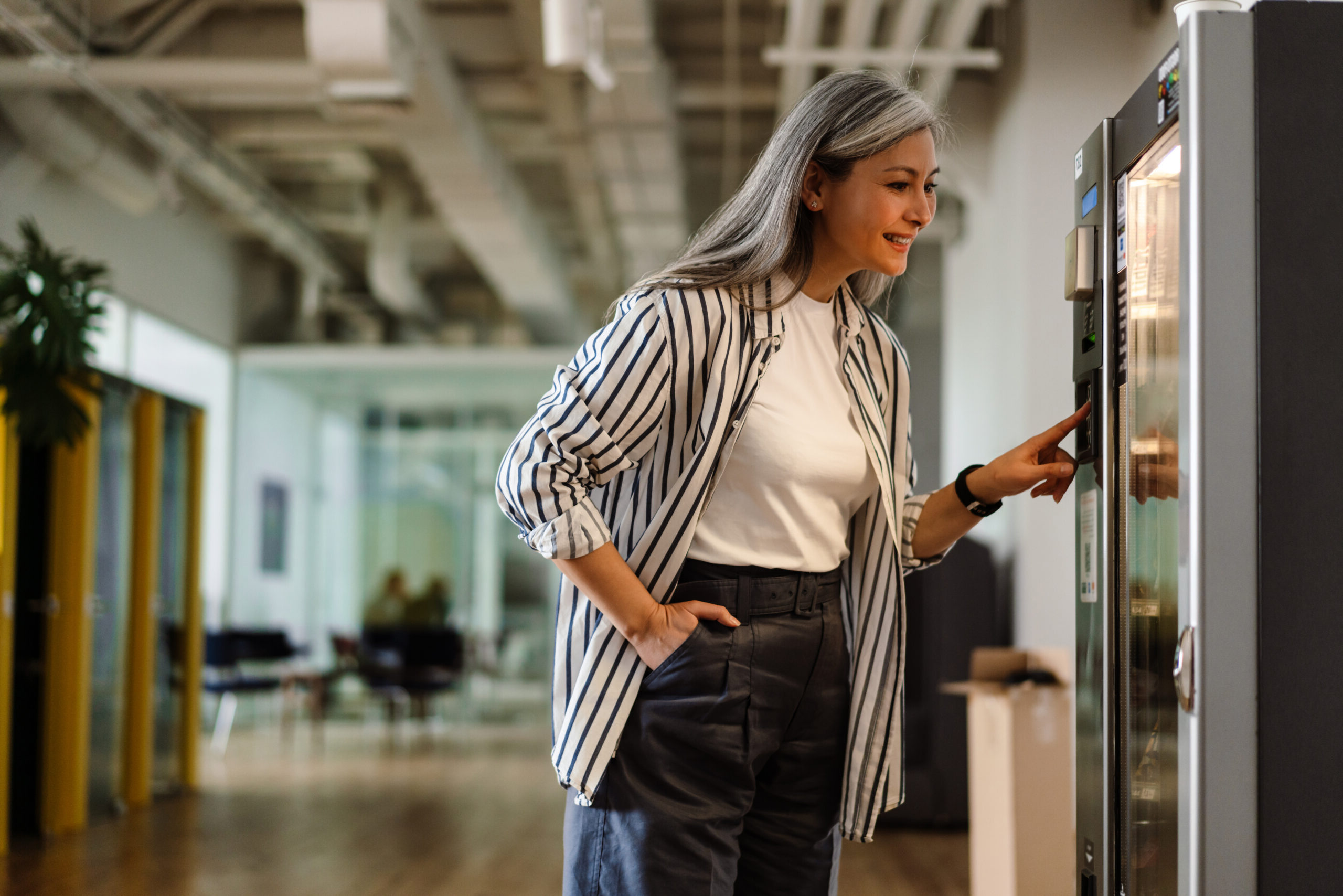 A woman with long gray hair browses a vending machine in a modern office space, smiling as she selects an item.
