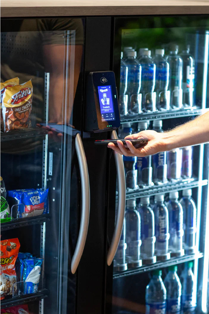A hand uses a digital payment device on a vending machine, which displays snacks and bottled drinks behind a glass door.